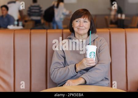 Valencia, Spain : March 18, 2023: Smiling middle aged woman in casual clothes holds a paper cup of hot Starbucks coffee with straw at Starbucks coffee Stock Photo