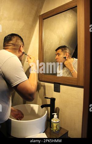 Forty-year-old dark-skinned Latino adult man shaves with a razor in front of the bathroom mirror at home because he is afraid of waxing Stock Photo