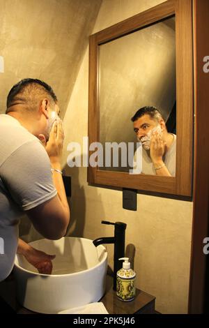 Forty-year-old dark-skinned Latino adult man shaves with a razor in front of the bathroom mirror at home because he is afraid of waxing Stock Photo