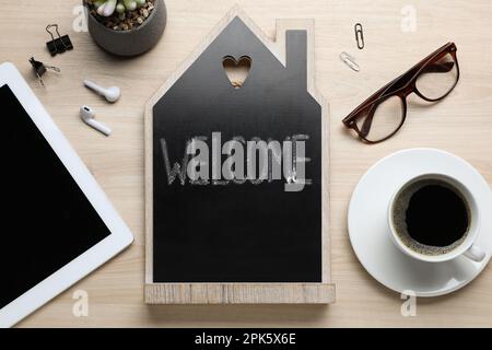 Flat lay composition of house shaped blackboard with word Welcome and gadgets on wooden table Stock Photo