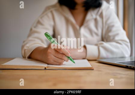 Close-up image of a young Asian female college student in a white hoodie writing something on a book, remote working at a coffee shop. Stock Photo