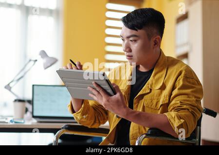 Young man with disability working in office of company, reading document on tablet Stock Photo