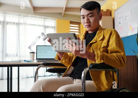 Serious businessman sitting in wheelchair and reading news on digital tablet Stock Photo