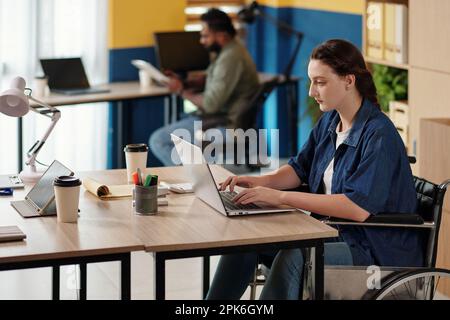 Serious young woman with disability working on laptop, answering e-mails from colleagues Stock Photo