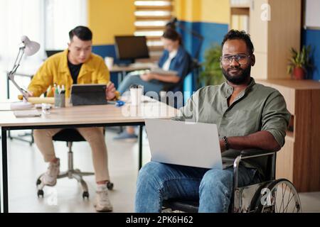 Portrait of positive software developer sitting in wheelchait with laptop and smiling at camera Stock Photo