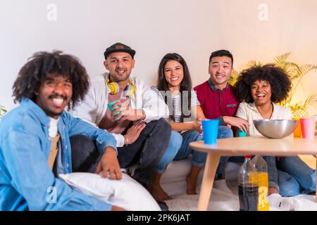 Group portrait of multiethnic friends eating popcorn and drinking soda on sofa. smiling looking at camera Stock Photo