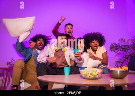 Adult party. Young people sitting on the sofa watching a football game with popcorn. Emotion for soccer, purple led, celebrating a goal Stock Photo