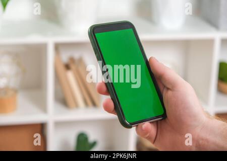 A man holds a phone with a green screen in his hands against a background of green house plants. Mockup. Chromakey. Stock Photo