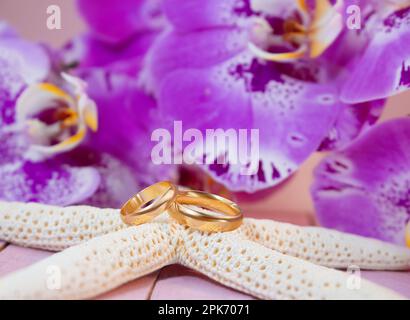 Golden wedding rings on white starfish behind purple orchids on pink wooden board. Celebrations, ceremonies, honeymoon Stock Photo