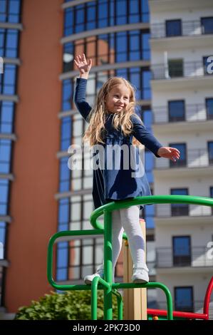 Cheerful girl climbing, holding hand up, hanging out at playground. Portrait of adorable female child playing on climbing frames, smiling in sunny day. Modern residential building on background. Stock Photo