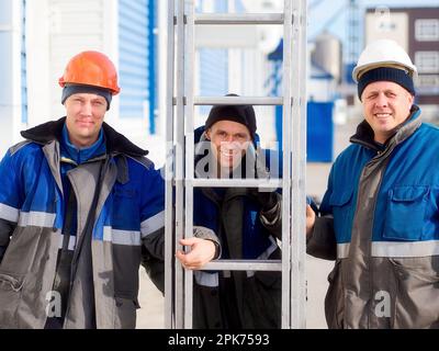 Three workers in winter overalls and helmets stand outside looking at camera and smiling. Real portrait of Caucasian laborers. There are happy builders with portable staircase outside.. Stock Photo