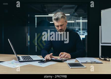 Serious thinking financier business analyst working with documents inside office, mature adult accountant on paper work calculating with calculator and laptop inside office. Stock Photo
