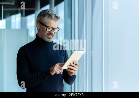Mature adult businessman standing near window inside office, man smiling and using tablet computer, gray-haired boss browsing internet pages and reading news. Stock Photo
