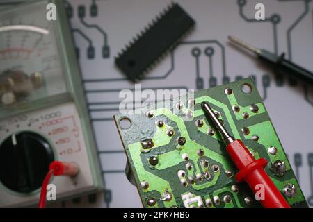 Electronic repairing bench. Electronic circuit board with blurred multimeter, chip and diagram background. Focused on red probe. Stock Photo