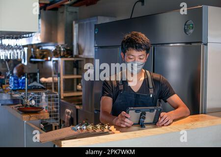 A man wearing a face mask at the counter of a restaurant kitchen, using a digital tablet, the owner or manager. Stock Photo