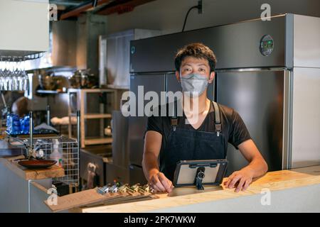 A man wearing a face mask at the counter of a restaurant kitchen, using a digital tablet, the owner or manager. Stock Photo