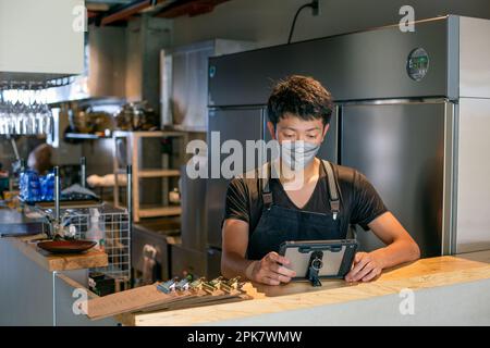 A man wearing a face mask at the counter of a restaurant kitchen, using a digital tablet, the owner or manager. Stock Photo
