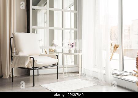 A white faux leather armchair near the window next to a small table with flowers. Part of the interior with copy space.High quality photo Stock Photo