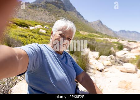 Happy senior biracial woman sitting on rock and taking selfie in mountains Stock Photo