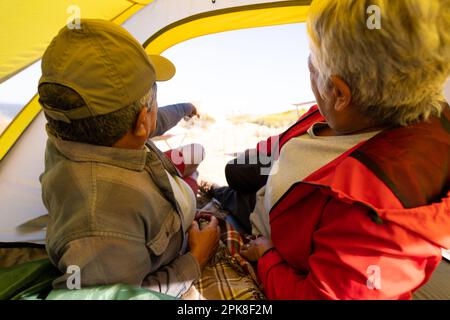 Happy senior biracial couple lying in tent and looking outside in mountains Stock Photo
