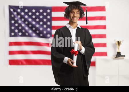 African american male student in a graduation gown holding a diploma and standing in front of a USA flag Stock Photo