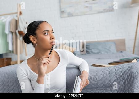 thoughtful african american student holding notebook and pen near books on bed,stock image Stock Photo