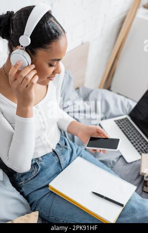 African american student in headphones holding cellphone near notebook on bed,stock image Stock Photo