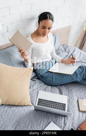 African american student in headphones holding book near laptop and notebook on bed,stock image Stock Photo