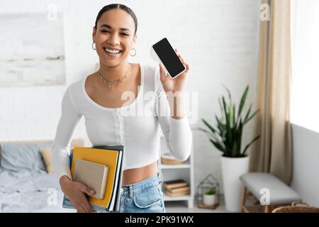Smiling african american student holding notebooks and showing cellphone in bedroom,stock image Stock Photo