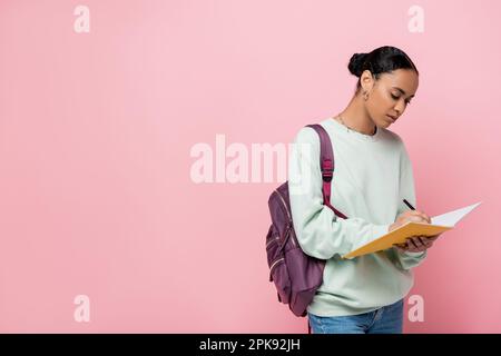 brunette african american student taking notes while holding notebook and pen isolated on pink,stock image Stock Photo