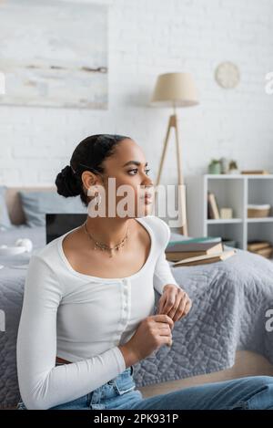 pensive african american student looking away while sitting near books on bed,stock image Stock Photo