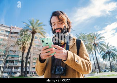 Happy bearded man using a cellphone app for browsing on internet. Tourist guy smiling and looking for location using a smart phone map standing outdoors. Male with eye glasses holding a mobilephone. High quality photo Stock Photo