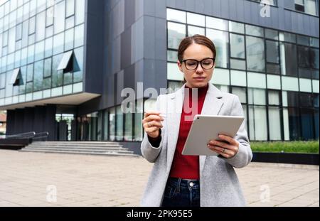 Businesswoman wearing eyeglasses working remotely on her digital tablet outdoors against business centre. Stock Photo