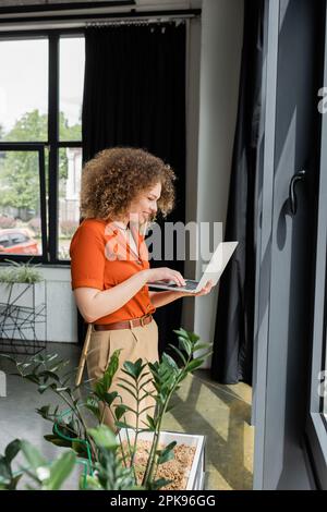 side view of happy businesswoman with curly hair using laptop next to green plants in modern office,stock image Stock Photo