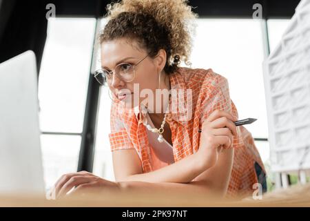 curly architectural designer using laptop while holding stylus near residential house model,stock image Stock Photo