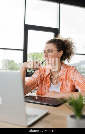 pensive architectural designer holding stylus pen near mouth while sitting near graphic tablet and laptop in office,stock image Stock Photo