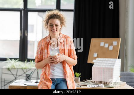 cheerful architectural designer holding paper cup while standing near house model in office,stock image Stock Photo