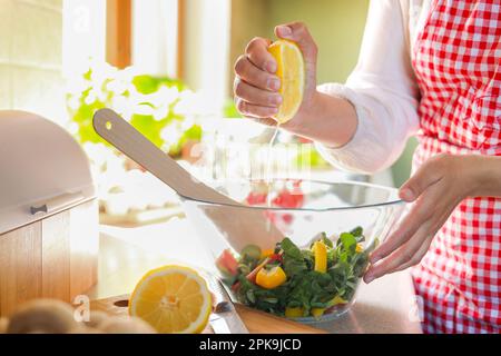 Woman squeezing fresh lemon over bowl with salad at countertop in kitchen, closeup Stock Photo