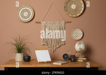 Stylish macrame and wicker wall decor hanging above wooden table with houseplants and stationery Stock Photo