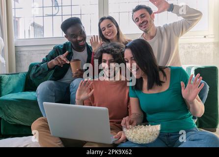 Five multi-ethnic millennial friends sitting on sofa looking at computer screen wave hands laughing enjoy distant communication with mate. Make videoc Stock Photo