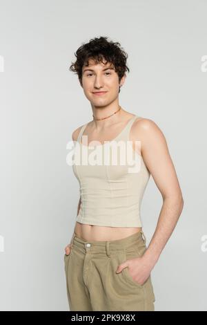 young bigender model in tank top and pants standing with hands behind ...