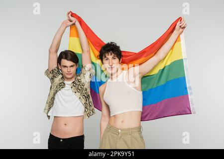 positive nonbinary couple in stylish clothes holding rainbow flag and looking at camera isolated on grey,stock image Stock Photo