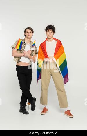 full length of young nonbinary models in stylish clothes posing with rainbow flags on grey background,stock image Stock Photo