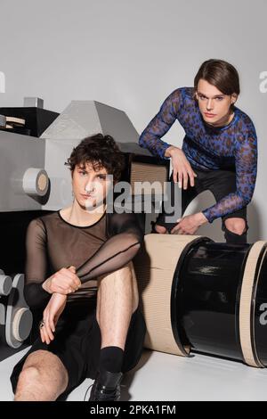 young pangender couple in fashionable tops posing near model of photo camera on grey background,stock image Stock Photo