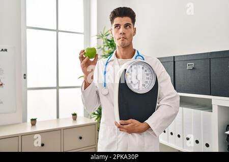 Young hispanic doctor man holding scale at dietitian clinic making fish face with mouth and squinting eyes, crazy and comical. Stock Photo