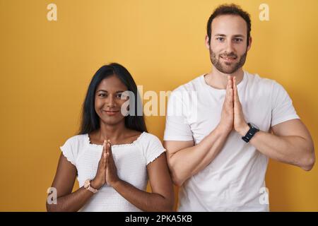 Interracial couple standing over yellow background praying with hands together asking for forgiveness smiling confident. Stock Photo