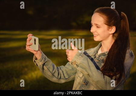 smiling teenager girl makes thumbs up gesture at smartphone camera in park, video call Stock Photo