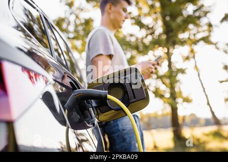 Young man charging electric car parked in the nature area and adjusting an EV charging app on smartphone Stock Photo