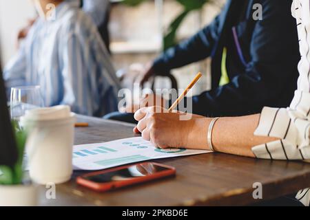 Close-up of woman's hand taking notes on chart paper in office meeting, wooden table with other people, papers, smartphone, and coffee cup. Stock Photo