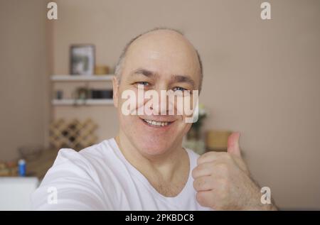 Smiling male blogger taking selfie or video call from his apartment, showing hand gesture. Stock Photo